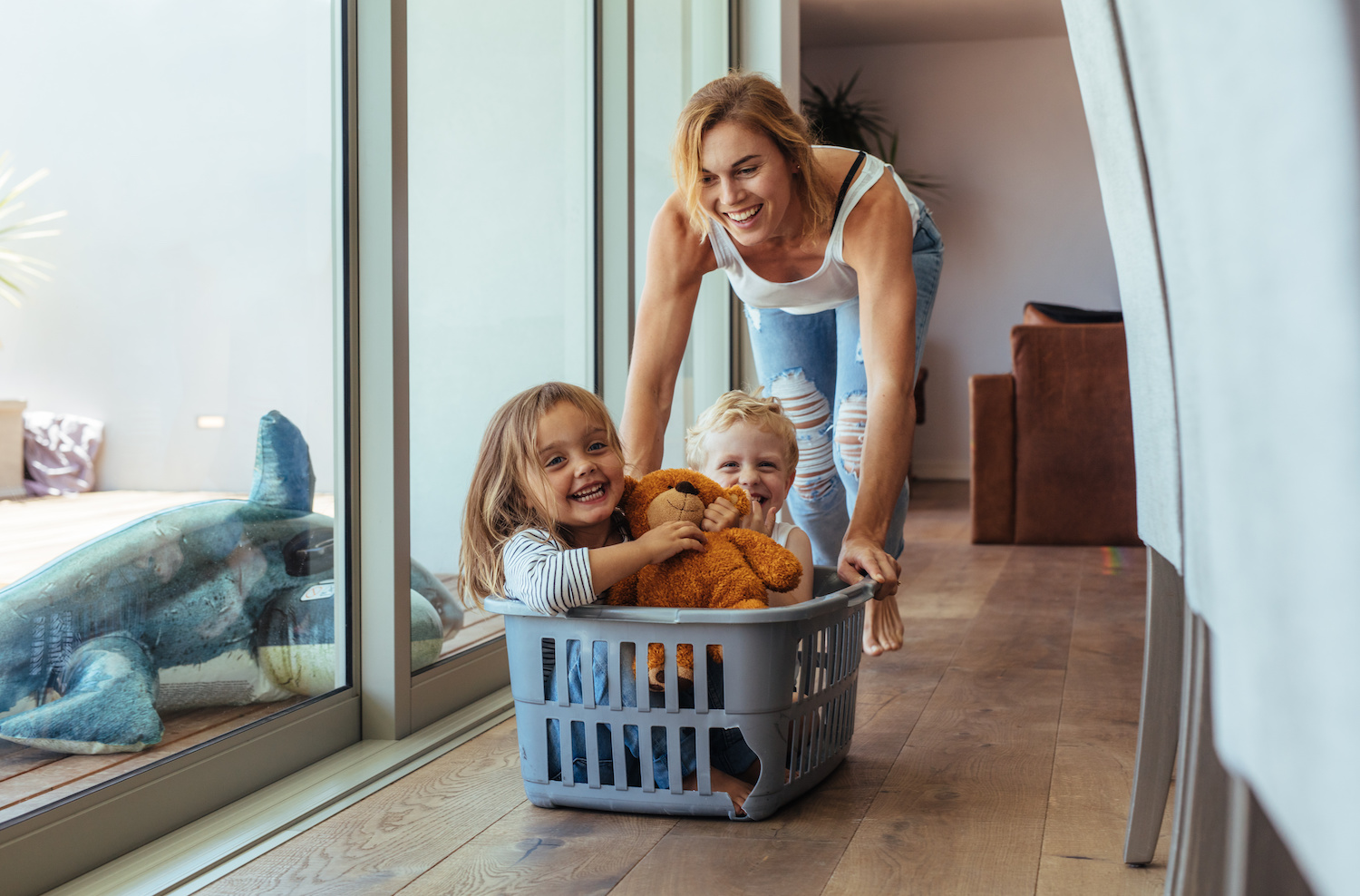 Happy young mother pushing children sitting in laundry basket. Mother and children playing at home.