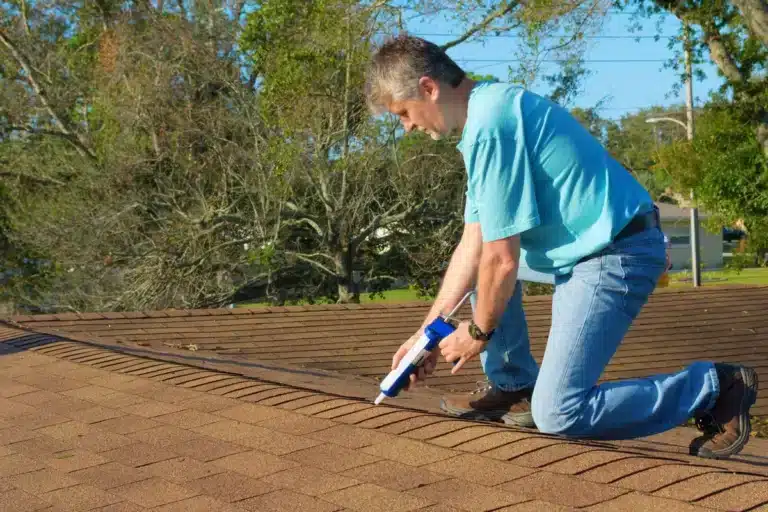A man in a blue shirt and jeans kneels on a shingled roof, applying sealant along the ridge with a caulking gun—demonstrating expert roof repair. Trees and power lines can be seen in the background.